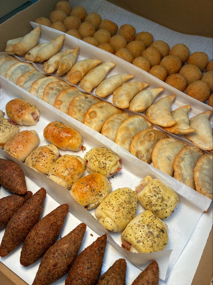A catering box filled with rows of various fried and baked appetizers, including kibbeh, empanadas, and cheese balls.
