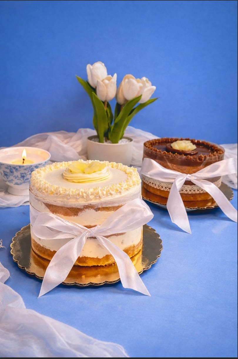 Two decorated cakes with white ribbons, a lit candle, and white tulips on a blue background.