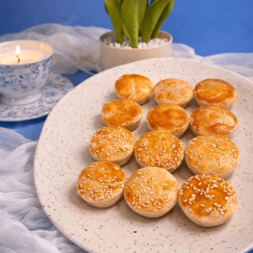 A plate of golden-brown mini pies, some topped with sesame seeds, next to a lit candle and plant.